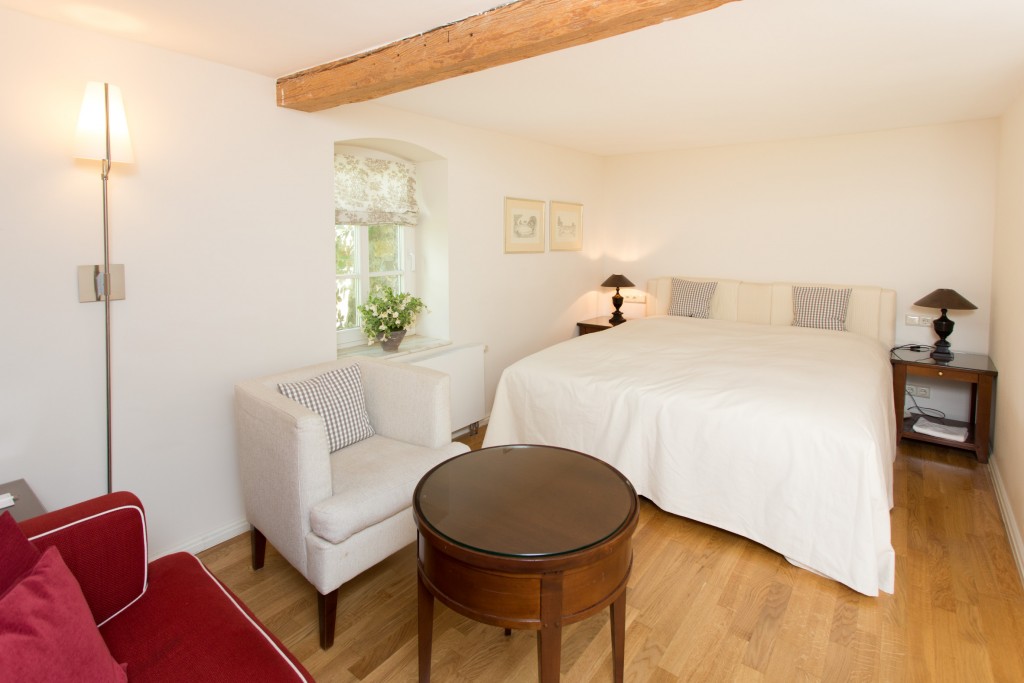 Helles Zimmer mit gemütlichem Doppelbett gegenüber einer roten Couch und einem weißem Sessel