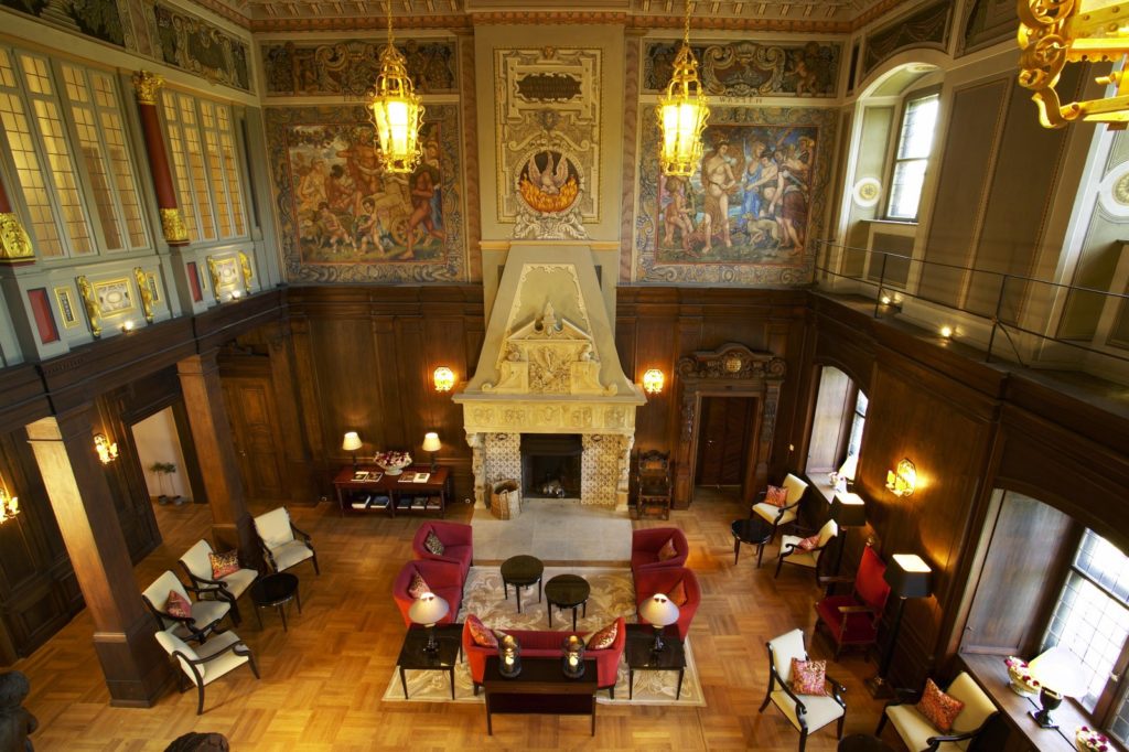 Historischer Rittersaal mit roten Sesseln an einem Kamin unter goldenen Laternen und bunten Gemälden
