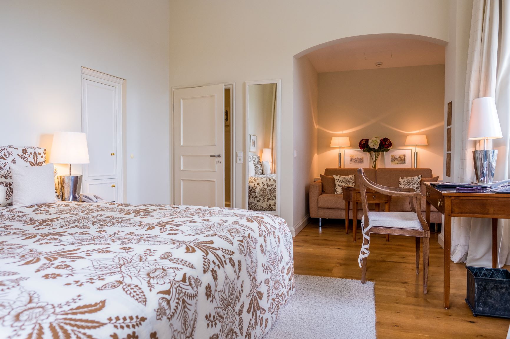 Großzügiges Zimmer mit gemustertem Doppelbett gegenüber eines Holzschreibtischen und einer Wohnnische mit brauner Couch