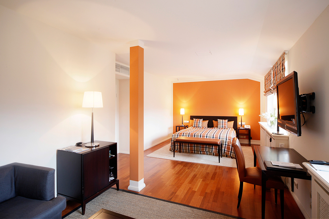 Kariertes Doppelbett vor orangener Akzentwand mit Schreibtischausstattung und grauer Couchgarnitur