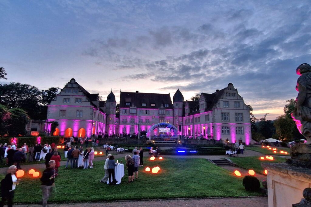 20240817_205900 Lila beleuchtetes Schloss an dämmrigen Abend mit Gästen auf dem Rasen davor